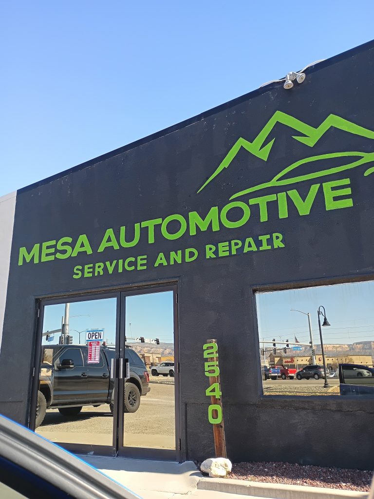 Exterior of an automotive service building with Mesa Automotive Service and Repair in bright green on a dark facade. A parked truck is reflected in the glass doors below an Open sign.