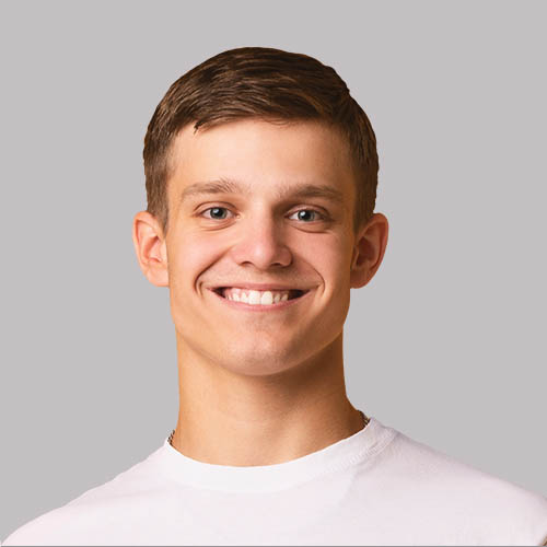 A young man with short brown hair and a bright smile wears a plain white t-shirt against a neutral gray background.
